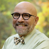 A person with a graying beard, round black glasses, a green shirt, and a yellow bowtie poses outdoors for a photo